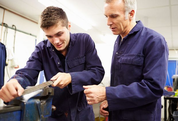 Older man watches younger man working on a vice